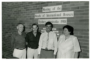Three business casual dressed men, one woman, smiling and standing in front of a brick wall with the signage "Meeting of the Heads of International Houses Sydney, Australia 1988"