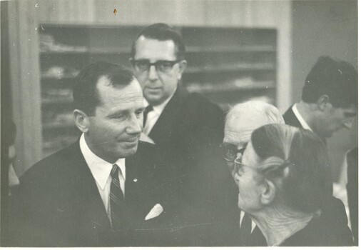 A low contrast photograph of a man in a suit in conversation with an older couple turned away from the camera. 