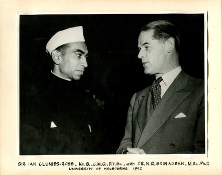A man standing with his arms crossed wearing a songkok looking at another man in a double-breasted suit. The man in the suit is speaking. 