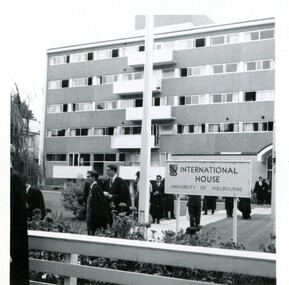 A five story building in the background, around ten male students in Oxford robes walking down the cement pathway.  A sign reads "International House. " 