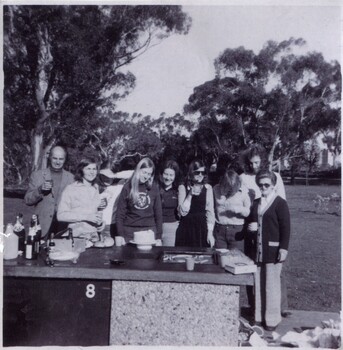 A group of people standing in front of an outdoor BBQ in a park. 