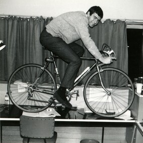 A male student in a dorm in riding pose on a bicycle on top of a desk. 