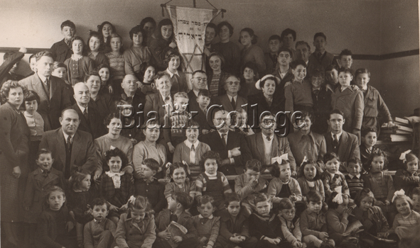 A black and white photo of a large group of people, including adults and children, seated and standing in rows facing the camera. In the middle of the back row is a large banner with Hebrew text and a Jewish star on it. 