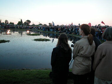 A crowd, some holding flags and banners, stand around a lake