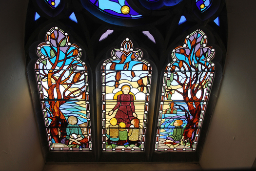 Detail of 3 pane stained glass window. Left and right panes have images of children under a tree.  Centre pane is of a teacher/parent with three children. May depict St Mary of the Cross