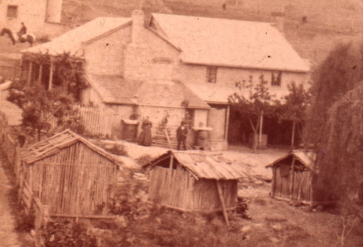 A woman and a man stand in front of a house, with 3 slab/wattle buildings in the foreground