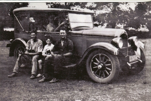 Three people sit on the running board of a vintage car
