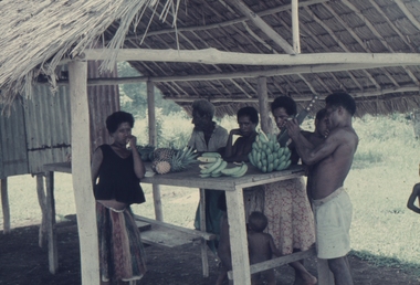 Papuans with bananas and other fruit in a shelter at Sogeri. Note the iron on the back wall.