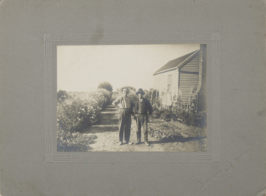 Image depicting 2 men standing in a garden, on a driveway, beside a house with chimney.