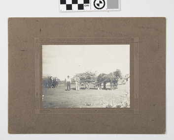 This image depicts 2 horse-drawn trailers loaded with cleared timber one man in background and 2 men in front of trailers on cleared ground