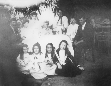 This image depicts a family of several generations seated around table older family members on chairs and standing 4 younger girls seated on ground two of which are holding dolls