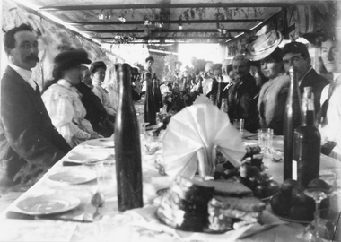 This image depicts a wedding celebration at long table with food and drink under a shelter, bride just visible on the left, women wearing hats and men in suits, at Merbein.