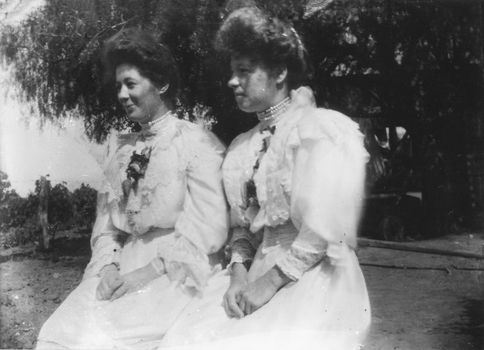 This image depicts 2 women sitting and dressed for a special occasion outside in a Merbein vineyard. Ladies dresses are a light colour trimmed in lace and wearing pearl chokers.