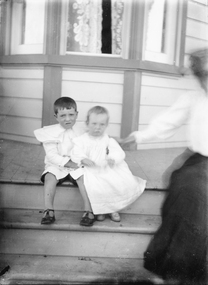 This image depicts two boys sitting on a verandah step, older child dressed in shorts and shirt with large collar and buckle up shoes, younger child wearing a long dress and covered shoes.