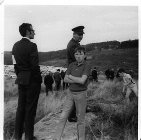 Photograph - VSSHP0310.04	047/A012	N0049	At Cardinia Reservoir's official works opening ceremony 1970		Black and white photo showing Greg Boothroyd, son of the photographer, looking at the camera as he stands on a low hill above the carpark. Behind him is a man in a peaked cap who is looking at him. Another man in a suit is watching proceedings. He has paper or similar tucked under his left arm. Another boy is running behind Greg. A large group of people is walking down a path to the left