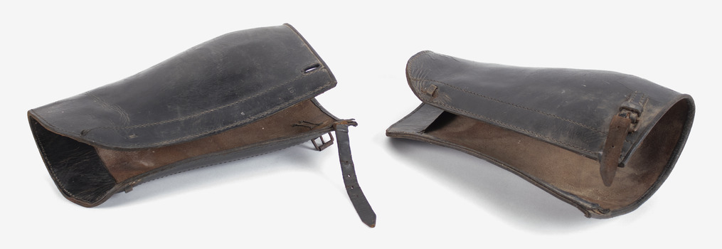 A pair of brown leather leg-gaters, lying on their side on a white back-ground. There is a buckle and strap to fasten at the top.