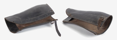 A pair of brown leather leg-gaters, lying on their side on a white back-ground. There is a buckle and strap to fasten at the top.