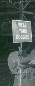 Photograph, "Wear Your Goggles"  - A safety sign on a workshop machine grinding stone from part of polio & tuberculosis (TB) patients undertaking occupational therapy & recreational activities in a workshop environment