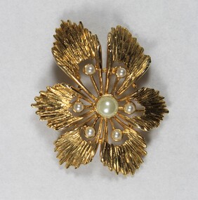 Front of a gold-toned metal brooch with one large central faux pearl surrounded by six smaller faux pearls.