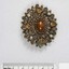 Front of an oval brooch from the Sarah Coventry jewellery range, with a central brown faux stone surrounded by four small faux pearls inlays and four small circular brown faceted glass inlays, and a 5 cm black and white scale athe bottom of the photo.