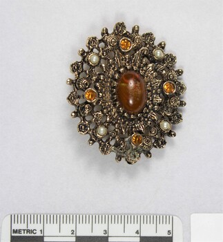Front of an oval brooch from the Sarah Coventry jewellery range, with a central brown faux stone surrounded by four small faux pearls inlays and four small circular brown faceted glass inlays, and a 5 cm black and white scale athe bottom of the photo.
