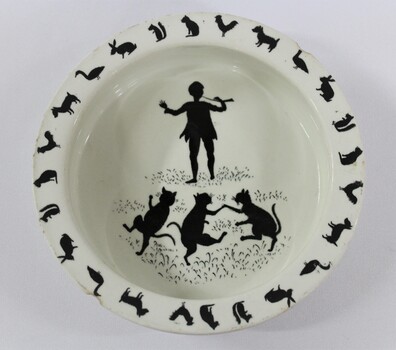 Birds eye view of a white ceramic child's bowl with black print depicting a child dancing with three cats in the centre of the bowl. A circular print around the edge of the bowl features animals including roosters, squirrels, dogs, cats, rabbits and geese