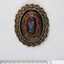 Front of an oval brooch in gold toned metal with red, blue and black inlay, with a 5 cm black and white scale.