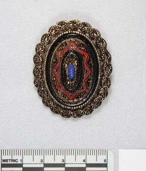 Front of an oval brooch in gold toned metal with red, blue and black inlay, with a 5 cm black and white scale.