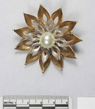 Front surface of a gold and silver toned metal brooch with a central faux pearl from the Sarah Coventry Pty. Ltd. jewellery range, with a 5 cm black and white scale.