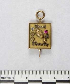 Front of a Sarah Coventry gold metal pin for ten years service, with a faceted pink stone inlay on the proper left side and a faceted clear stone on the proper right side, and a 5 cm black and white scale.