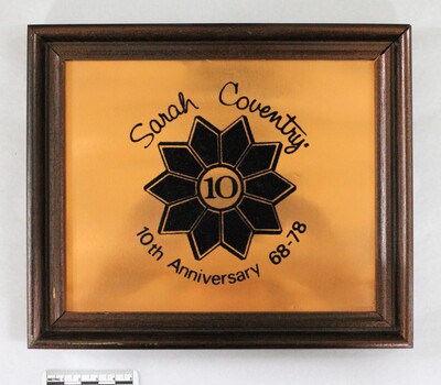 Front of a brown wooden fame on a copper plaque commemorating Sarah Coventry 10th Anniversary from 68-78, with a 5 cm black and white scale.