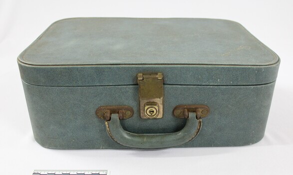 Front of a green suitcase with a handle and a metal lock for a Sarah Coventry jewellery demonstration kit and a 5 cm black and white scale.