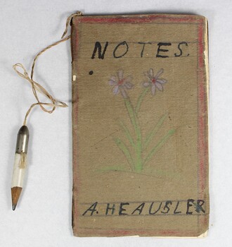 A handmade child's notebook with small white pencil attached by a piece of string. A hand drawn purple flower is on the front cover of the notebook, and the pages are blank.  