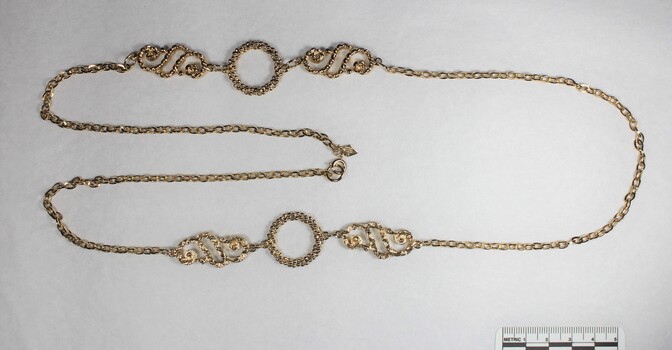 Gold toned metal chain with two circular elements and two swirling elements, with a black and white 5 cm scale.