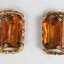 The front of two gold toned metal clip-on earrings with rectangular brown glass settings.