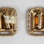 The back of two gold toned metal clip-on earrings with rectangular brown glass settings, and a label attached with "U.S.A. visible"