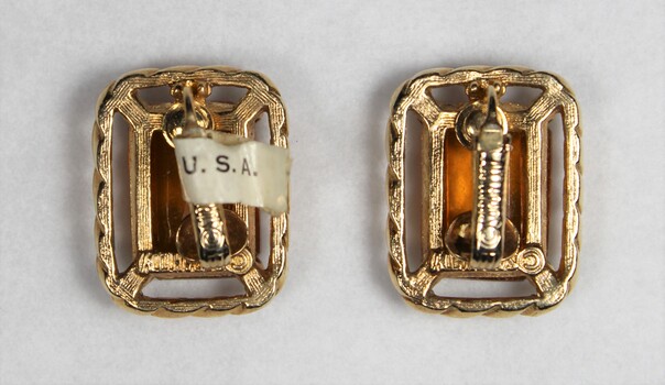 The back of two gold toned metal clip-on earrings with rectangular brown glass settings, and a label attached with "U.S.A. visible"
