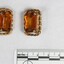 The front of two gold toned metal clip-on earrings with rectangular brown glass settings and a black and white 5 cm scale.