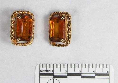 The front of two gold toned metal clip-on earrings with rectangular brown glass settings and a black and white 5 cm scale.