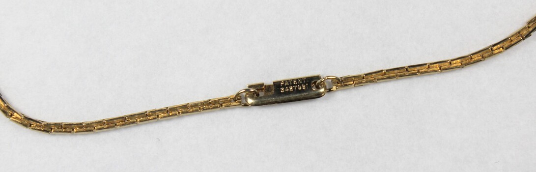 Detail of the back of the clasp on the gold toned metal chain with the oval-shaped pendant with the patent number.