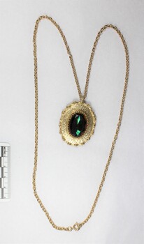 Front of a gold toned metal cable chain necklace with an oval pendant and a faceted green/brown glass setting and a black and white 5 cm scale.