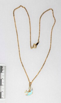 Front of a necklace with a gold toned metal chain and a small blue bird pendant and a small faux pearl, with a black and white 5 cm scale.