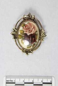Front of a gold toned metal brooch-pendant with a cameo of one pink and one red rose, with a black and white 5 cm scale.