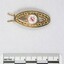 Front of a gold toned metal hair clip with a circular enamel inlay and a black and white 5 cm scale. 