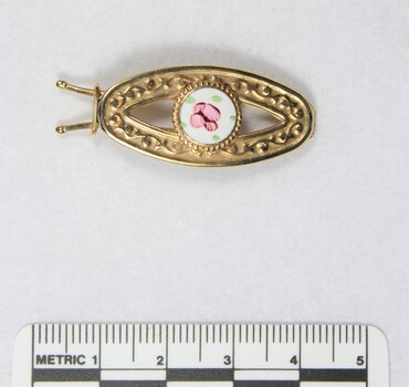 Front of a gold toned metal hair clip with a circular enamel inlay and a black and white 5 cm scale. 