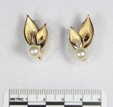 Front of two gold toned metal earrings with each earring consisting of a faux pearl and two leaf-shaped elements, and a black and white 5 cm scale.