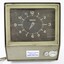 Closer view of the front of the green metal time clock with a brown clock face and light coloured numbers. Two labels attached to the lower front surface with a horizontal lock between them and electrical cords extending from one side. 