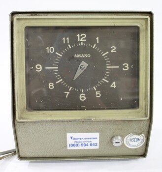 Closer view of the front of the green metal time clock with a brown clock face and light coloured numbers. Two labels attached to the lower front surface with a horizontal lock between them and electrical cords extending from one side. 