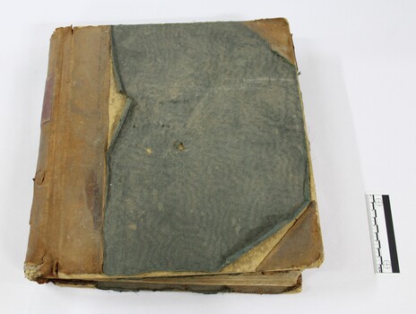 Front of a bound ledger with a deteriorated light brown leather and green textile cover, with a black and white 5 cm scale.