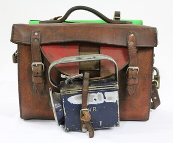 Front of a Victorian Railways guard's satchel, with a dark blue metal lamp attached to the front and rolled up green signal flag attached to the top.